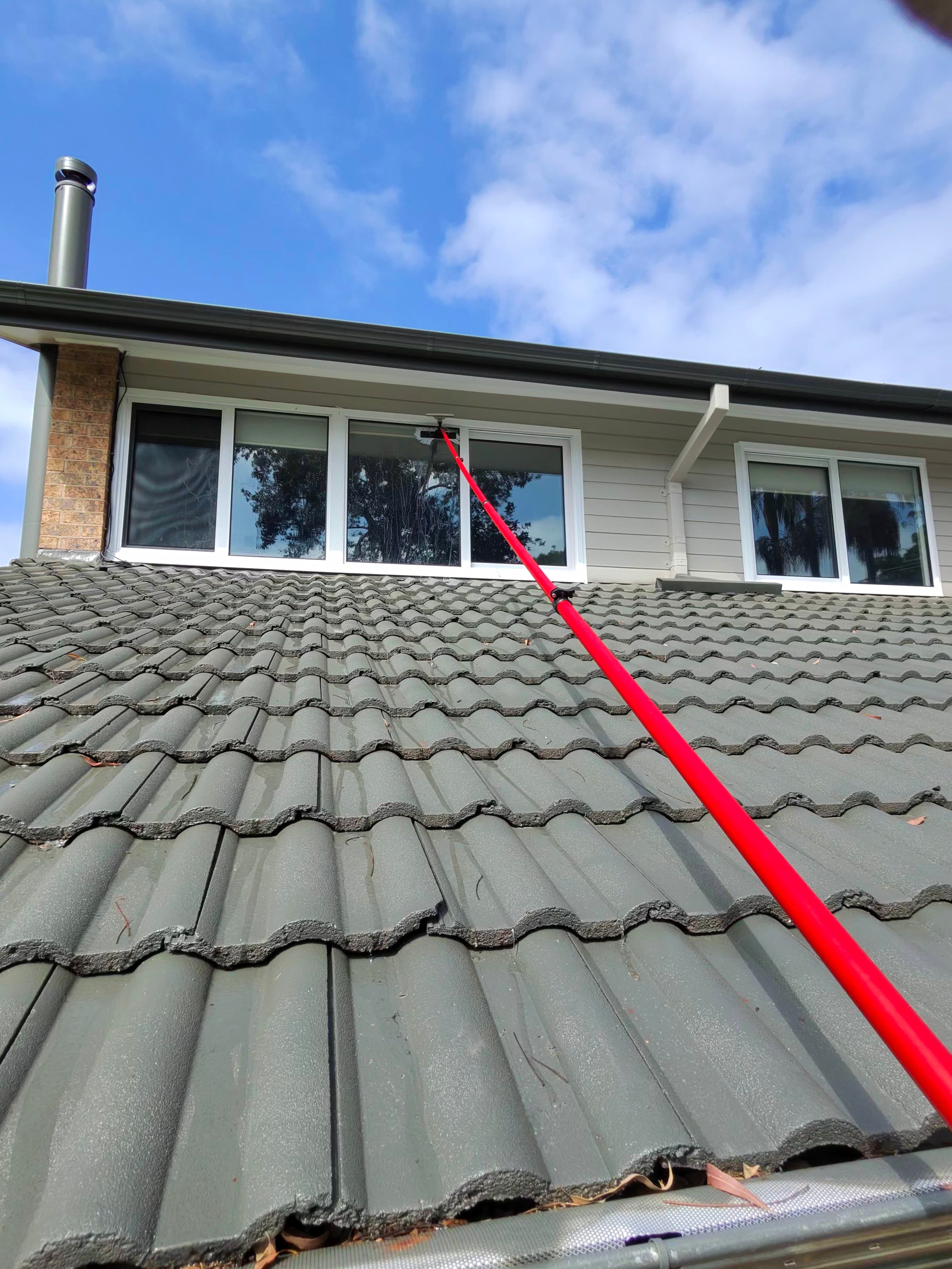 Long red telescopic pole cleaning second-story windows from a grey tiled roof.