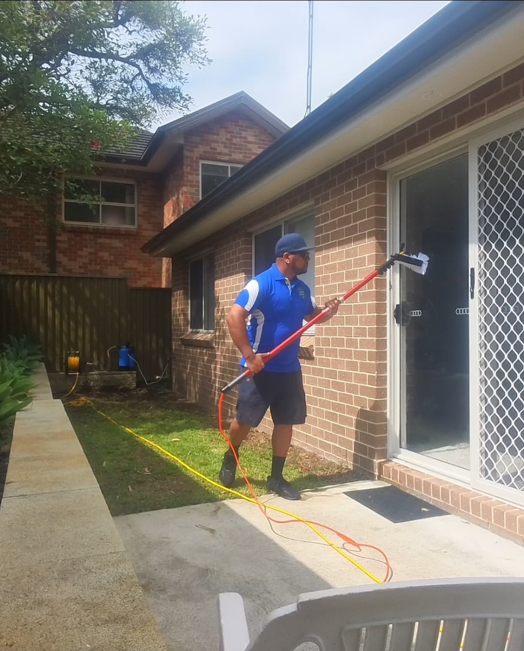 Man in blue shirt cleaning a glass sliding door with a long-handled brush outside.