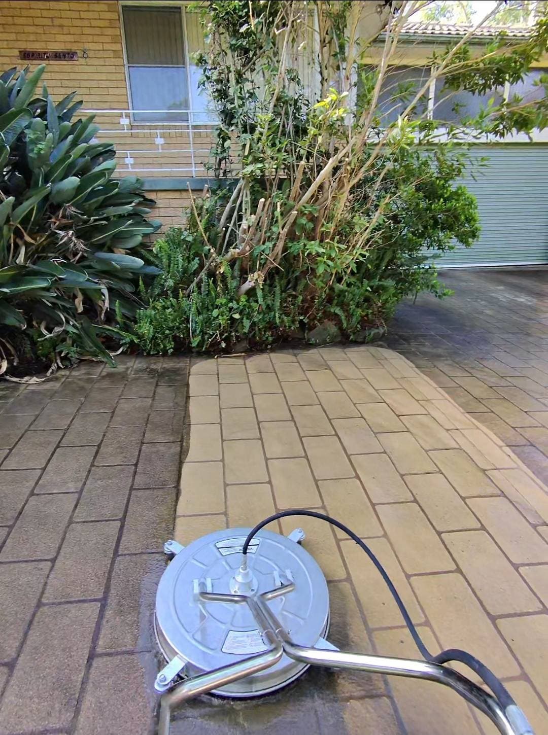 Surface cleaner pressure washing a brick driveway, revealing clean tan pavers next to dirty ones.