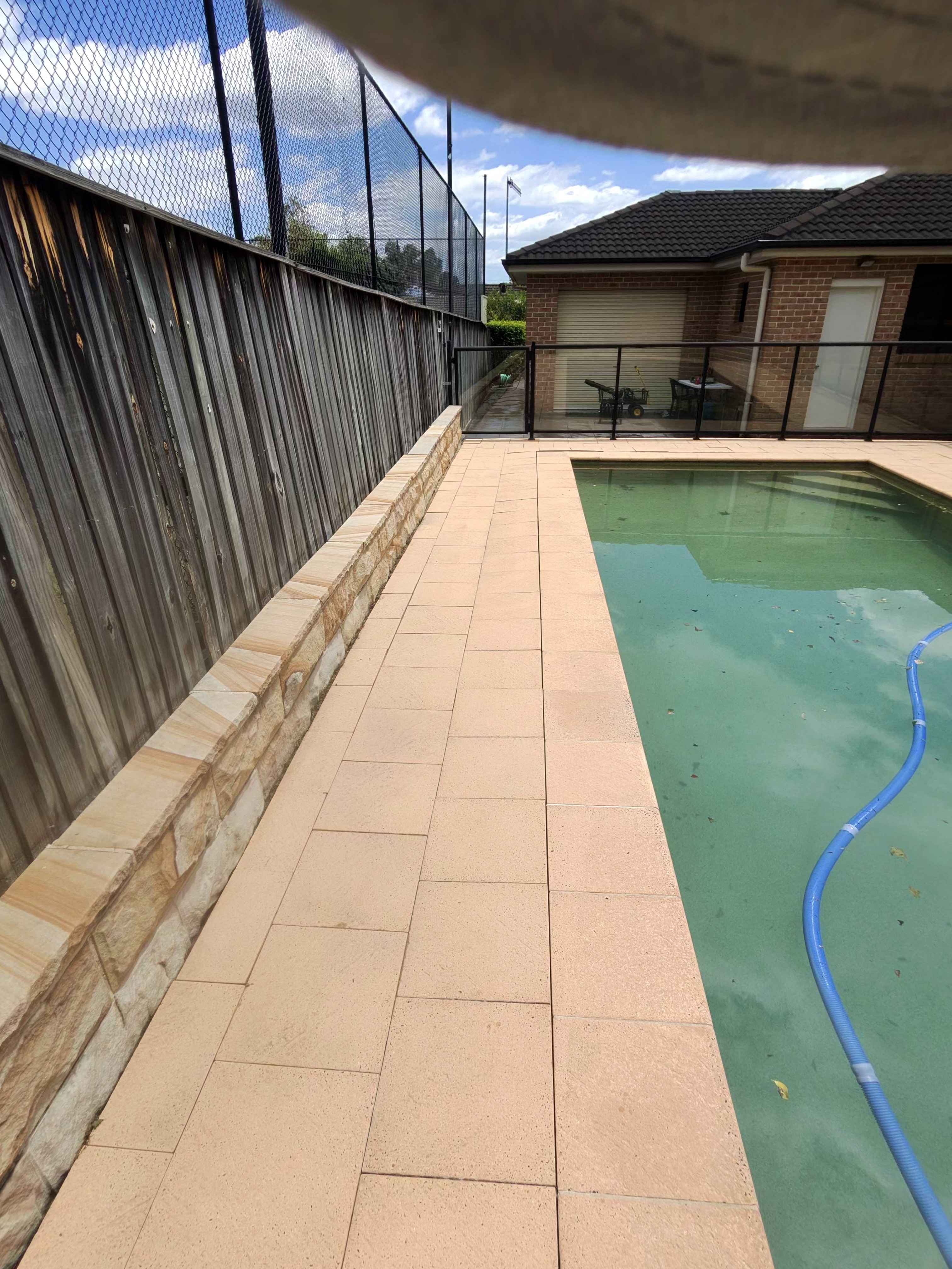 Backyard swimming pool area featuring tan pavers, a low stone wall, and a high wooden fence.