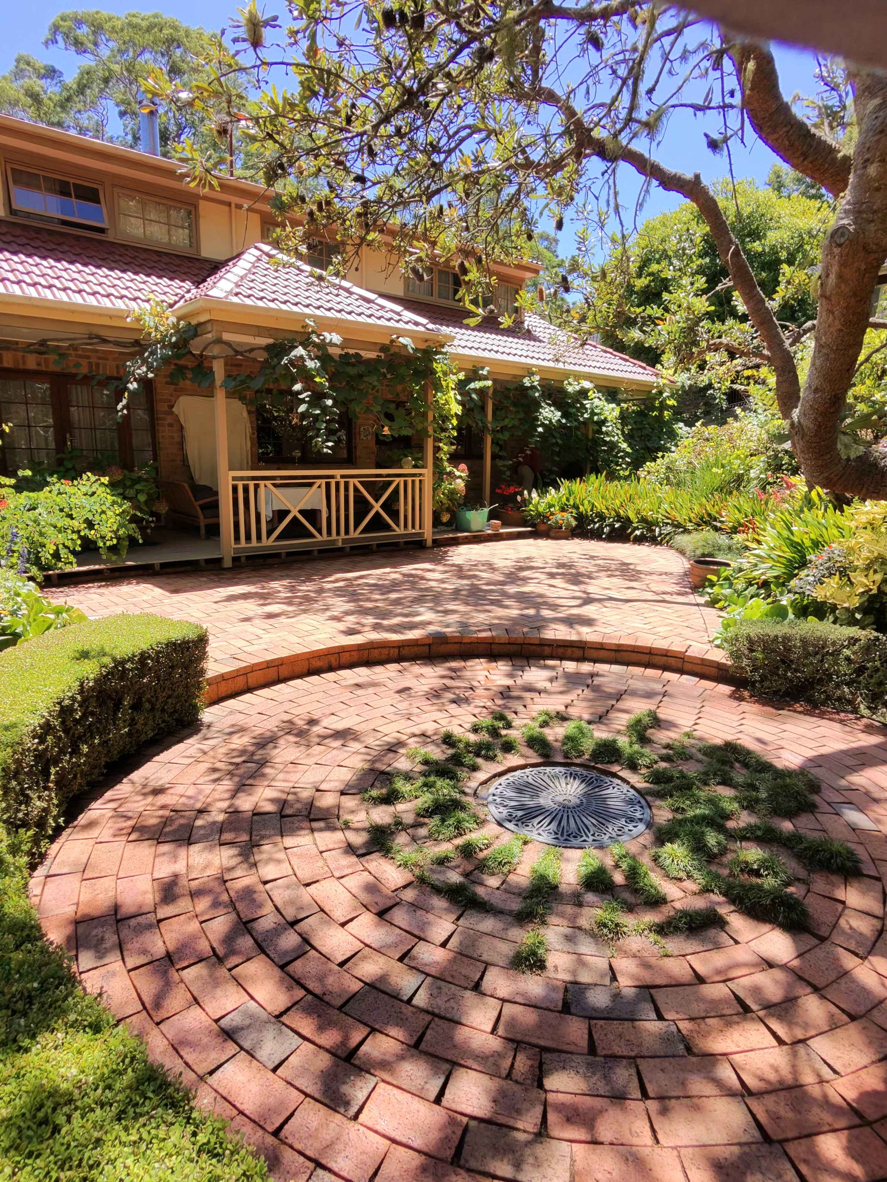 Circular brick patio with a decorative grate, surrounded by lush greenery and a red-roofed house.