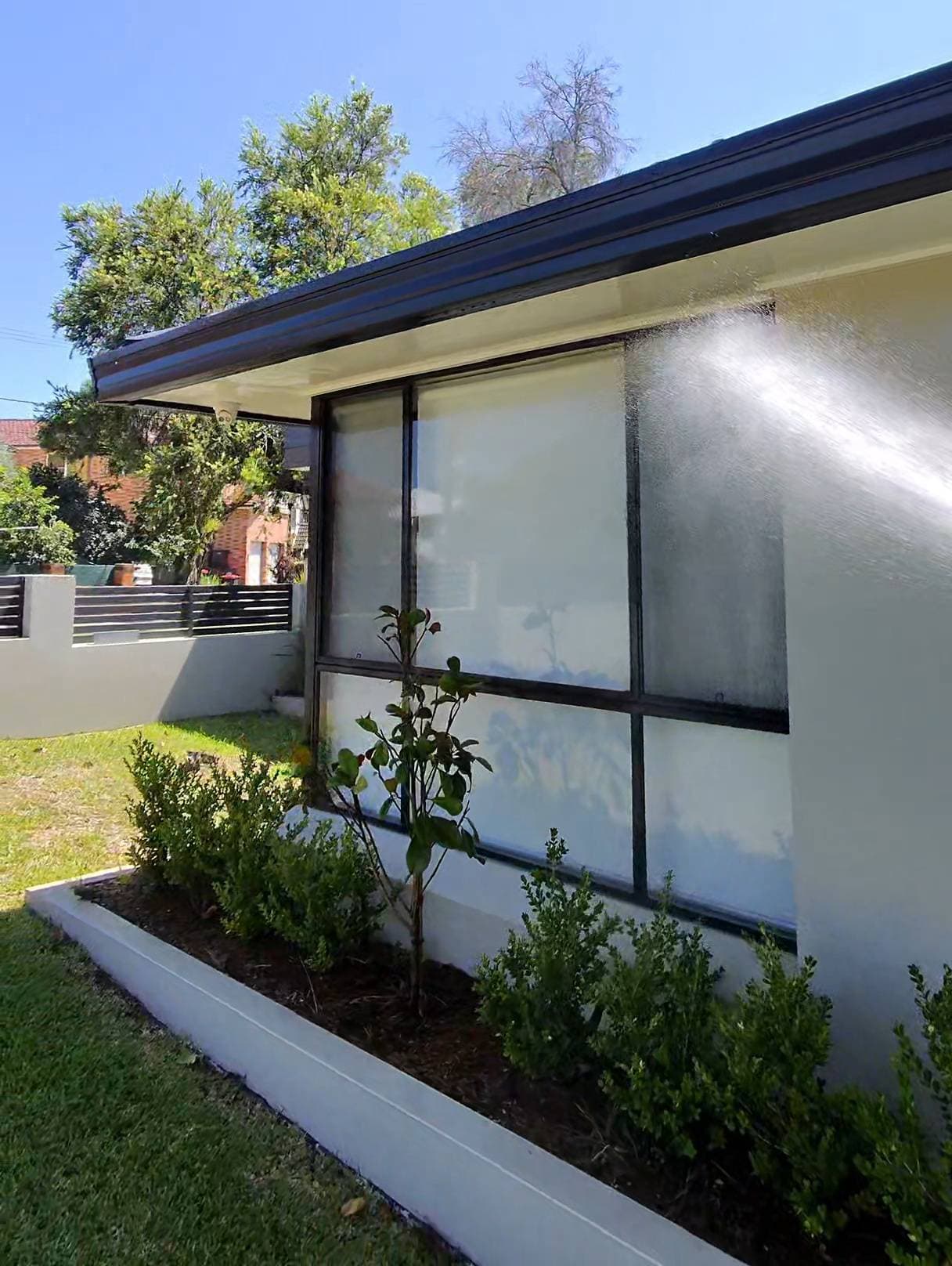 Water sprays against a white house wall and large window above a small garden bed.